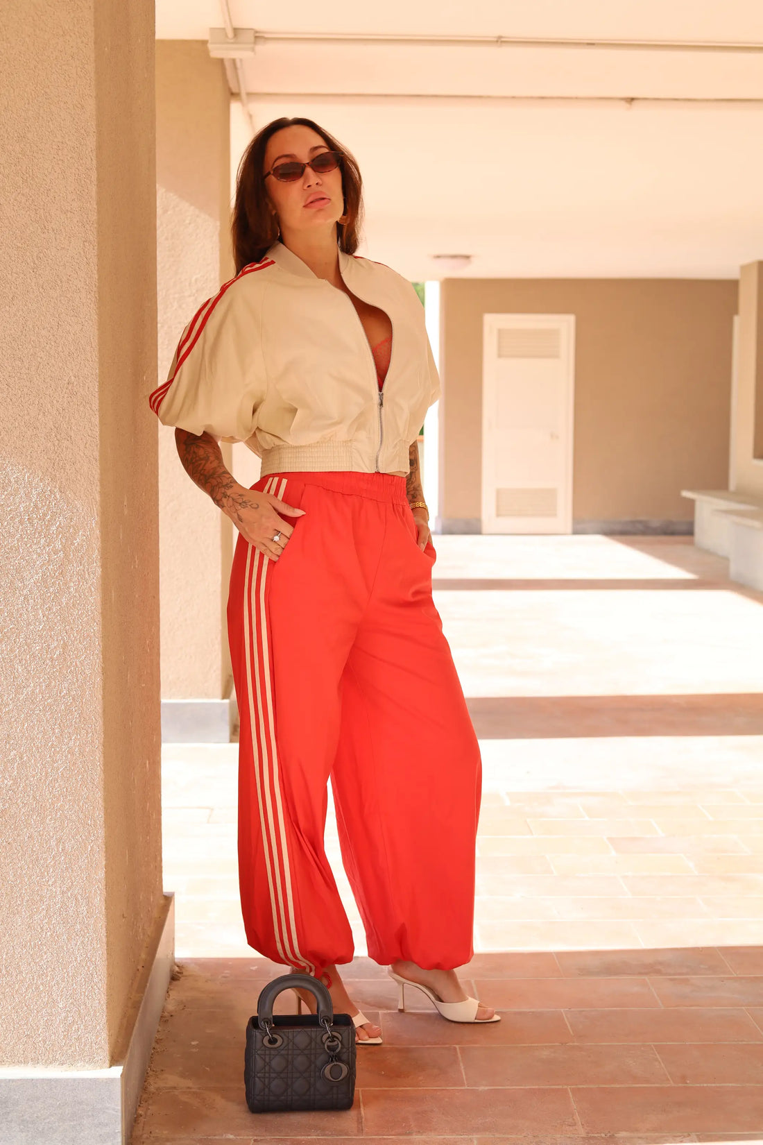 Woman in beige jacket and red pants standing in a sunlit hallway.
